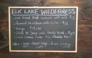 Chalkboard. Text: Elk Lake Wilderness, Hot Roast Beef Sandwich with side $15, Peameal on a bun with side $12, Wings $14 per pound, Chili or soup with garlic toast $8.50, Chili Cheese Fries - $12, Poutine $10, Sides: Soup, Chili, Onion Rings (+ $2), Fries, Poutine (+$3).